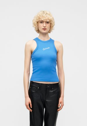 Jeune personne aux cheveux blonds courts et bouclés, portant un haut bleu sans manches avec le texte « Gelato » et un pantalon en cuir noir, debout devant un fond blanc.