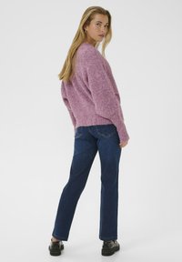 Femeie cu părul lung și blond, purtând un pulover roz pufoș, blugi de culoare albastră închisă și încălțăminte neagră, stând și privind peste umăr.