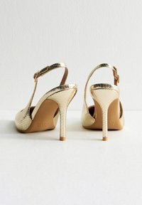 Guld, präglade slingbackklackar med hög stilettklack. Har en texturerad yta och en justerbar spännestraps.