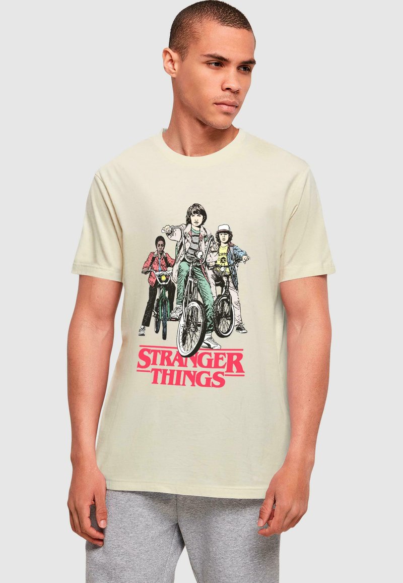 Cremefarvet bomulds t-shirt med et grafisk tryk af tre figurer på cykler med "STRANGER THINGS" i rødt nedenunder.