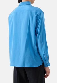 Camicia a maniche lunghe azzurro chiaro con colletto a camicia, piega sul retro e polsini con bottoni. Realizzata in tessuto leggero e liscio.