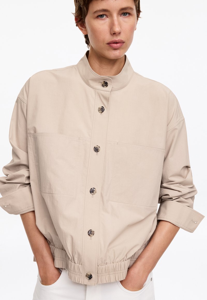 Personne portant une veste beige boutonnée avec un col montant et une taille élastique, mains dans les poches, sur fond blanc.