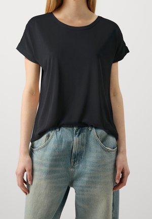 Vrouw draagt een losse zwarte korte mouwen shirt, lichtjes ingestopt in een lichtblauwe high-waisted spijkerbroek, met haar handen langs haar zijden rustend.