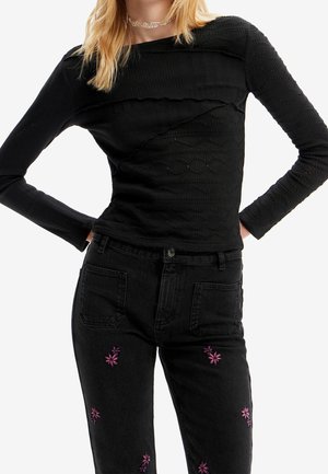 Schwarzes, geripptes Langarmoberteil mit strukturierten Paneelen, kombiniert mit schwarzen Jeans, die lila Blumenstickereien an den Beinen aufweisen.
