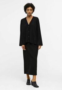 Zwarte lange mouwen cardigan met een V-hals en 5 knopen, gecombineerd met een zwarte enkellange rok en zwarte sleehakken. Zachte textuur.