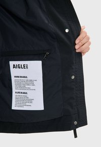 Intérieur de veste imperméable noire avec une étiquette blanche contenant du texte, une poche zippée et des fermetures à pression le long du bord. Texture lisse.
