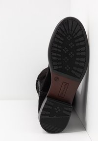 Suela de bota negra con dibujo de textura, tamaño 37, y marca Tamaris, fotografiada sobre una superficie blanca en una esquina.