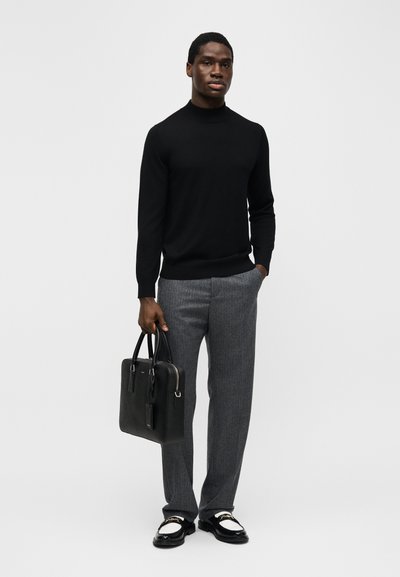 Άνδρας που φοράει μαύρο πουλόβερ ζιβάγκο, γκρι παντελόνια, μαύρα και λευκά μοκασίνια, κρατώντας μαύρη δερμάτινη τσάντα ενάντια σε απλό φόντο.