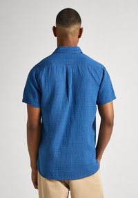 Camisa azul de manga corta hecha de tejido texturizado, con yugo trasero y dobladillo curvado. Diseño informal sin patrones o acentos visibles.
