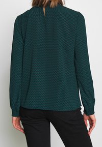Donna che indossa una blusa fantasia verde scuro a maniche lunghe e pantaloni neri, vista di schiena su uno sfondo bianco uniforme.