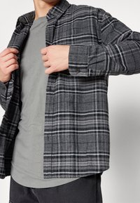 Camisa de franela a cuadros en gris oscuro con líneas blancas; mangas largas y puños con botón; llevada sobre una camiseta gris, mostrando su textura suave.