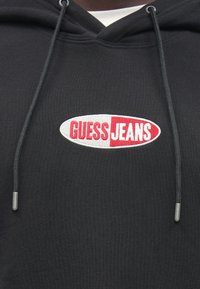 Μαύρο φούτερ με κουκούλα με κεντημένο λογότυπο "GUESS JEANS" σε κόκκινες και λευκές αποχρώσεις. Διαθέτει κορδόνια και μεταλλικά άκρα. Απαλή υφή.