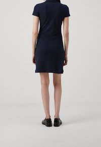 Nainen seisoo selin, päällään lyhythihainen tummansininen mekko ja mustat loafertossut, yksinkertaista harmaata taustaa vasten.