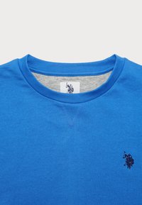 Blå crewneck sweatshirt med liten mörkblå broderad polo-spelare logotyp på bröstet och matchande etikett inuti kragen.