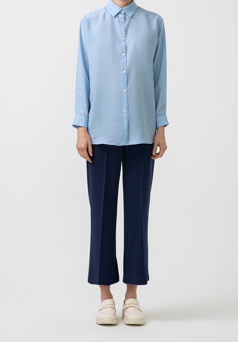 Personne portant une chemise bleu clair boutonnée, un pantalon large court bleu marine et des chaussures sans lacets blanc cassé, debout devant un fond uni.