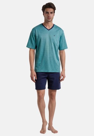 Jonge man die blootsvoets staat, draagt een tealkleurig T-shirt met korte mouwen en V-hals met borstzakje en marineblauwe korte broek tegen een effen achtergrond.