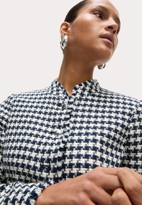 Giacca in tweed a quadretti blu navy e bianca con bottoni argentati e colletto alla mandarino, caratterizzata da tessuto testurizzato e dettagli di cucitura visibili.