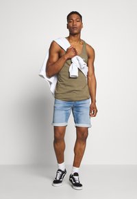 Olivgrünes Tanktop, hellblaue Denim-Shorts mit aufgeschlagenen Säumen, weiße Sneakers und ein weißes Sweatshirt über der Schulter drapiert.