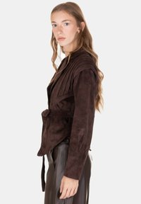 Giacca in camoscio marrone con design plissettato, dettaglio con cintura e maniche lunghe. Indossata sopra pantaloni neri in pelle, caratterizzati da una silhouette aderente.