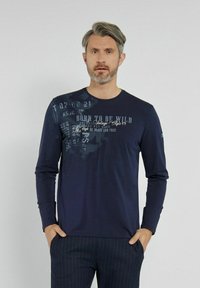 Granatowa koszulka z długim rękawem z nadrukowanymi tekstami w jasnoszarym i kremowym kolorze. Miękka tkanina i okrągły dekolt, w połączeniu z ciemnymi spodniami w paski.