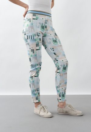 Persoană purtând pantaloni mulati cu model geometric în culori pastelate și pantofi casual bej cu șireturi, stând lângă un fundal simplu.