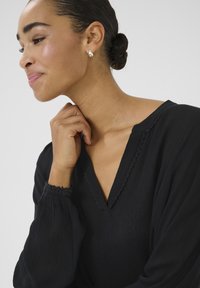 Black, textured blouse with a V-neckline and scalloped trim, featuring puffed long sleeves. Model wears teardrop earrings and has a bun hairstyle.