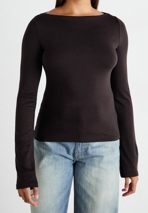 Kobieta w dopasowanym czarnym topie z długim rękawem i dekoltem w łódkę oraz jasnoniebieskich dżinsach, na tle jednolitego tła.