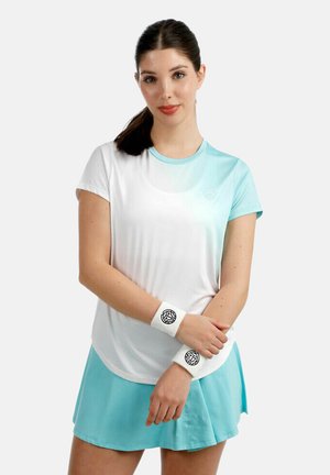 Jeune femme en tenue de sport blanche et bleu clair, portant des bracelets et une jupe, debout les bras croisés devant un fond uni.