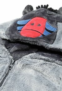 Grå plyschhuva med en broderad röd ansikte och blåa ränder. Har dragkedjestängning och texturerat fleece-material. Djurliknande öronaccenter på huvan.