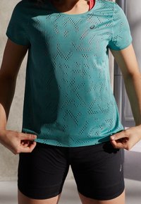 Turkusowa koszulka sportowa z perforowanymi wzorami i krótkimi rękawami, zestawiona z czarnymi, teksturowanymi szortami. Posiada logo w lewym górnym rogu.