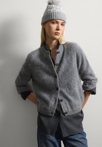 Cárdigan de lana gris con una textura suave, cierre de botones y mangas enrolladas, superpuesto sobre una camisa oscura. El modelo lleva un gorro de punto gris a juego.