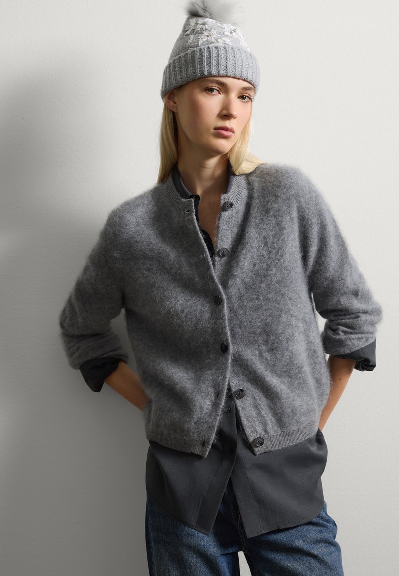 Cárdigan de lana gris con una textura suave, cierre de botones y mangas enrolladas, superpuesto sobre una camisa oscura. El modelo lleva un gorro de punto gris a juego.