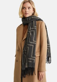 Auklas un vilnas maisījuma šalle tumši pelēkā krāsā ar gaišām ģeometriskām apdrukām un pušķotiem galiem, uzlikta uz brūnas mēteles. Mīksta tekstūra, taisnstūra forma.