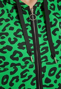 Felpa verde con zip e stampa leopardata nera, dotata di una zip argento e lacci neri, realizzata in tessuto a coste con tasche anteriori.