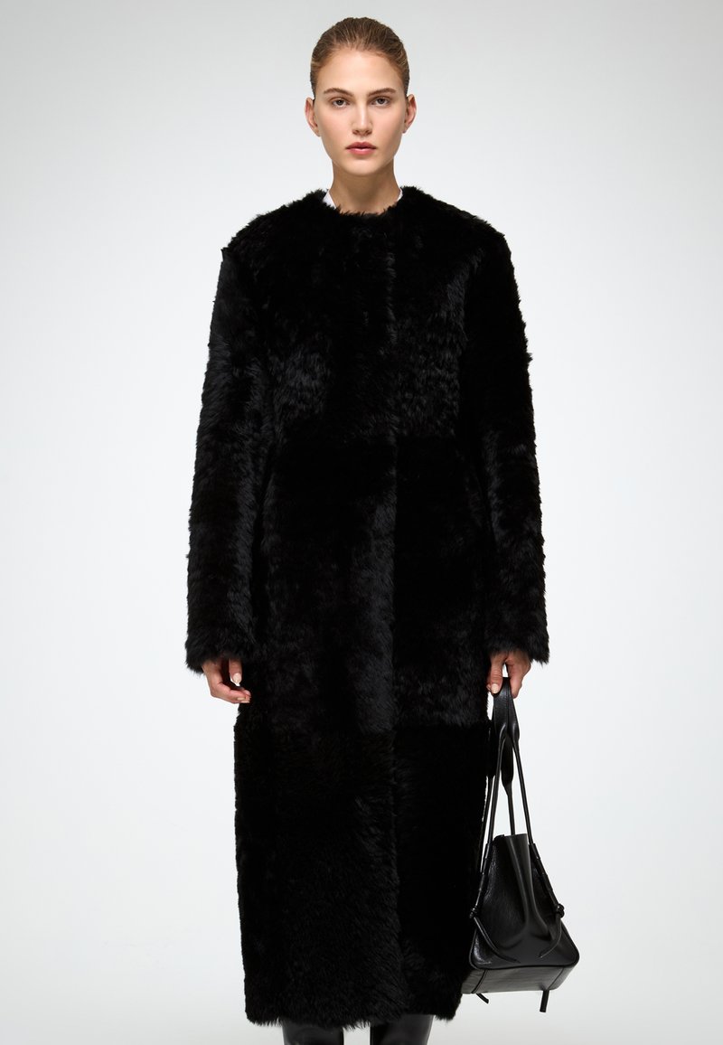 Cappotto in pelliccia sintetica nera con design dritto, maniche lunghe e lunghezza fino al polpaccio. Dotato di una texture liscia e senza chiusure visibili.