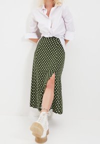 Jupe midi verte à pois avec une fente latérale ; associée à une chemise blanche nouée et des bottes de combat blanches. Texture lisse et coupe ajustée.