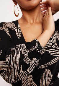 Czarna sukienka z materiału z beżowymi abstrakcyjnymi wzorami. Posiada głęboki dekolt w kształcie litery V oraz bufiaste rękawy z marszczeniami i detalem w postaci guzika przy mankiecie.