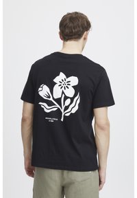 Sort bomuld t-shirt med et stort hvidt blomsterdesign på ryggen, med tekst under grafikken. Korte ærmer og rund halsudskæring.