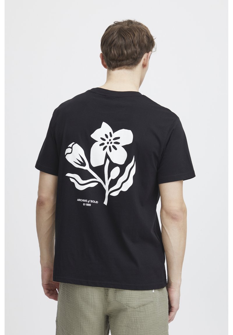 Sort bomuld t-shirt med et stort hvidt blomsterdesign på ryggen, med tekst under grafikken. Korte ærmer og rund halsudskæring.