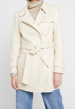 Kobieta w kremowym wełnianym płaszczu z paskiem, noszącym na beżowym golfie i niebieskich dżinsach, stojąca na jednolitym tle.