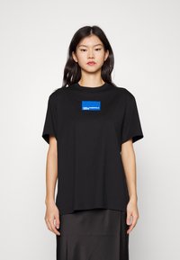 Camiseta de algodón negra de manga corta, con un logotipo rectangular azul en el pecho. Corte simple y recto, con una textura suave.