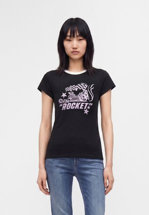 Jeune femme aux cheveux noirs portant un T-shirt noir avec "The Rocket" et un graphique de voiture, assorti à un jean bleu, debout devant un fond uni.
