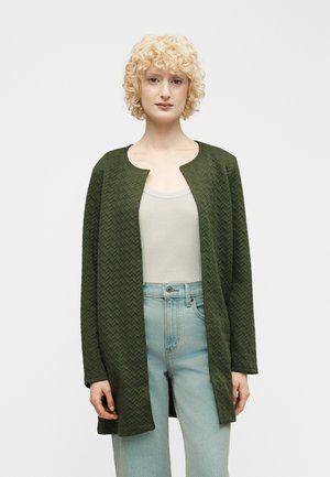 Blondine met een open, gestructureerde donkergroene lange mouw jas over een licht tanktop en lichtblauwe high-waisted jeans.