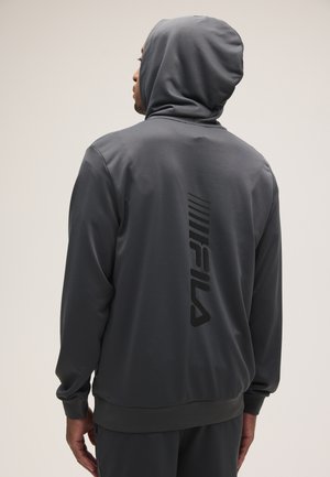 Hombre con sudadera con capucha gris oscuro y capucha puesta, de espaldas a la cámara mostrando el logo vertical de FILA en la espalda.