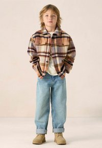 Flanellen jas in een bruin, oranje en crèmekleurig ruitpatroon met twee borstzakken, gecombineerd met lichtblauwe spijkerbroek en beige schoenen.