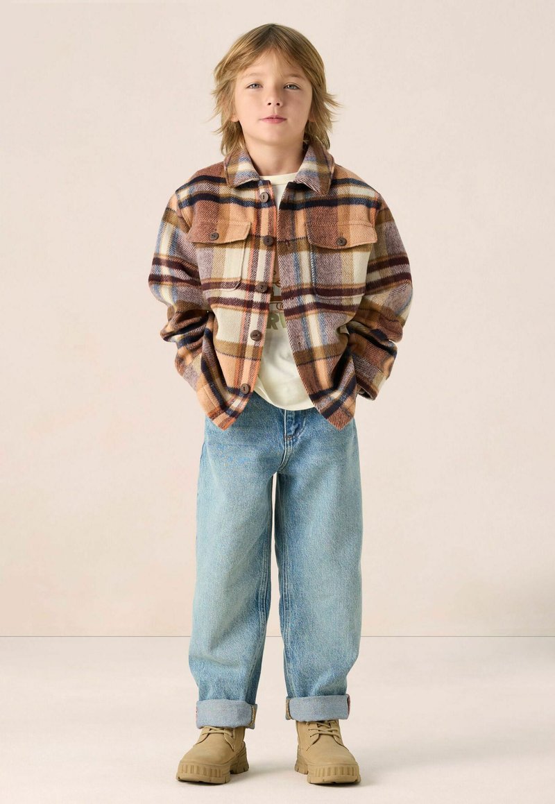 Flanellen jas in een bruin, oranje en crèmekleurig ruitpatroon met twee borstzakken, gecombineerd met lichtblauwe spijkerbroek en beige schoenen.