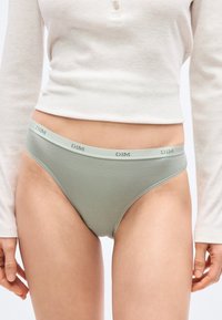 Γυναίκα που φοράει ανοιχτό πράσινο σλιπ ψηλής κοπής με το λογότυπο "DIM" στη μέση και εκρού μακρυμάνικο κοντό τοπ, στέκεται με χαλαρά τα χέρια.