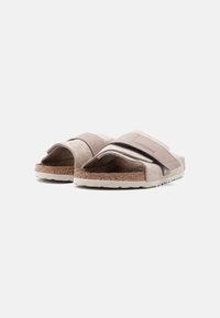 Ανοιχτό μπεζ σουέντ σαγιονάρες Birkenstock με ρυθμιζόμενους ιμάντες και πάτους από φελλό σε λευκό φόντο.