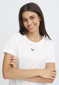 Fehér pamut póló rövid ujjakkal, három apró fekete szívmintával a mellkasán. Sima textúra és klasszikus fazon.