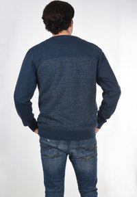 Marineblaues Sweatshirt mit strukturiertem oberen Rücken, der ein dezentes, horizontales Muster aufweist; gerippte Bündchen und Saum. Getragen mit blauen Jeans.